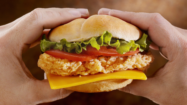 Closeup of hands holding a fried chicken sandwich with lettuce, tomato, and cheese