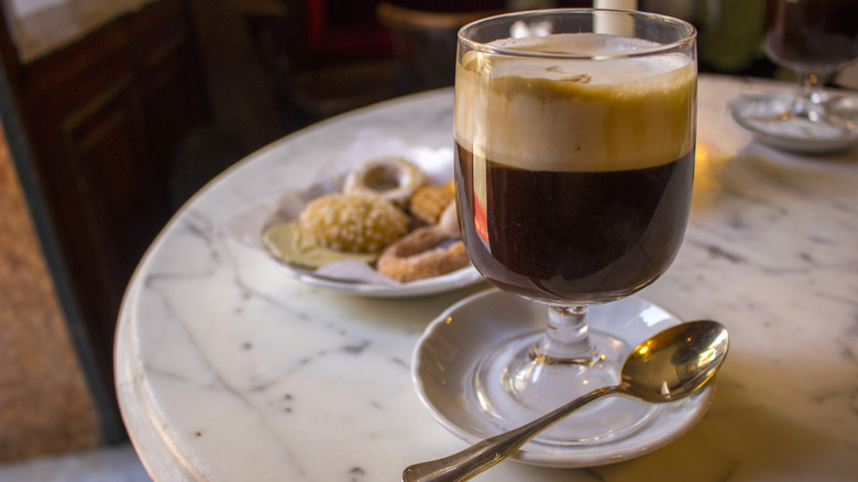 Clear glass of bicerin with layers of espresso, hot chocolate, and cream on top with cookies on the side at a cafe.