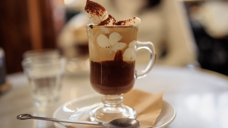 Bicerin glass with layered coffee, chocolate, and cream on top, sprinkled with cocoa powder, all on a plate with a spoon on the side and blurred cafe in background