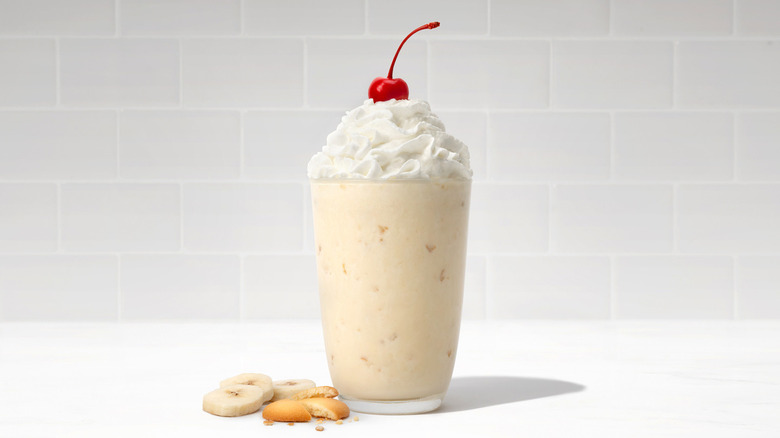 Chick-fil-A banana milkshake topped with whipped cream and a cherry next to some banana slicers and vanilla wafers against a gray brick wall