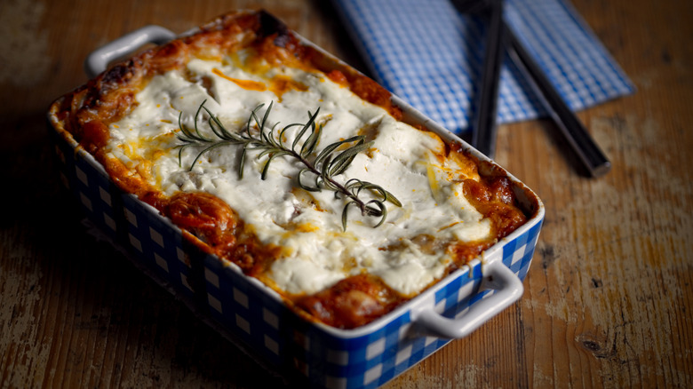 Fully cooked lasagna in a blue and white checkered deep pan, on a wooden table with utensils and a napkin in the background.