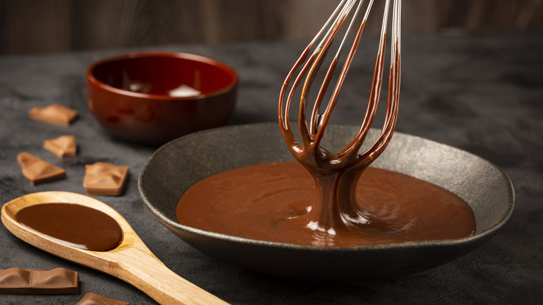 A whisk drips a liquid chocolate mixture into a bowl, with a wooden spoon filled with more of the mixture and broken bits of solid chocolate on the counter adjacent