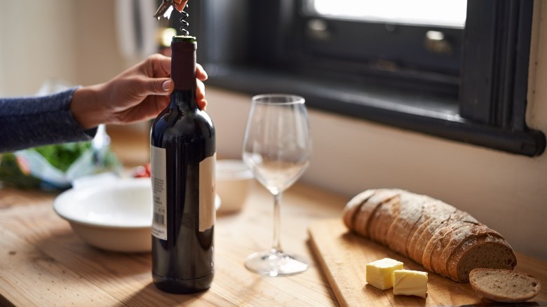 Person opening a bottle of wine on a wooden counter with bread 