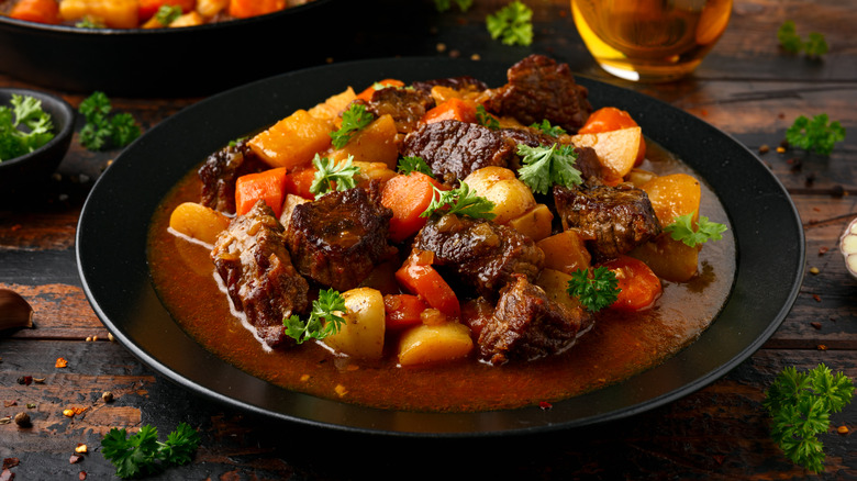 A bowl of beef stew on a countertop with potatoes and carrots, topped with parsley and surrounded by parsley sprigs, peppercorns, and chile flakes.