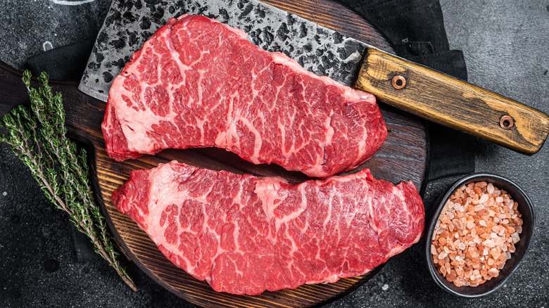 Two marbled, raw Denver cut steaks on a platter with a butcher knife underneath one, surrounded by thyme sprigs and salt.