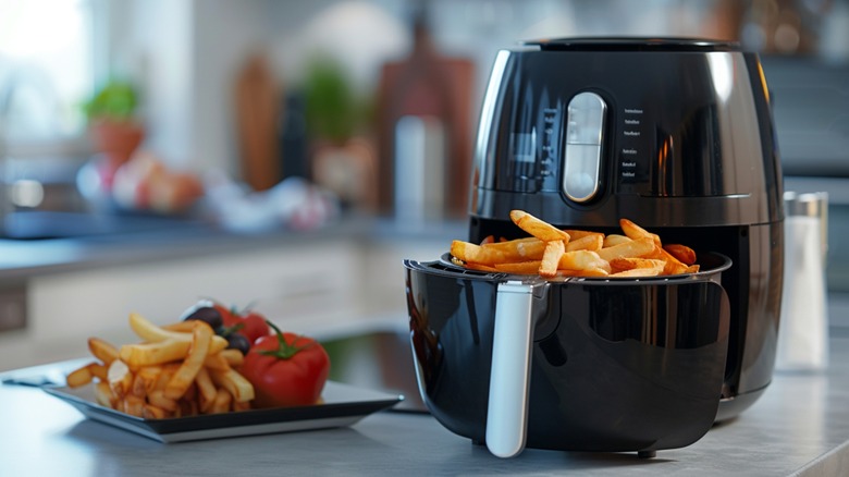 A black air fryer filled with french fries next to a plate with more fries and red bell peppers