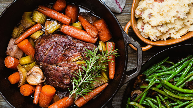 Pot roast in cast iron dutch oven with vegetables next to dishes of mashed potatoes and green beans