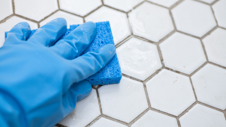 A hand in a blue glove uses a blue sponge to clean a white tile floor