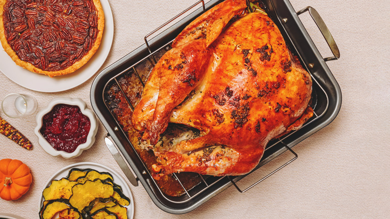 A cooked turkey sits in a roasting pan on a table aside cranberry sauce, pecan pie, and roasted vegetables