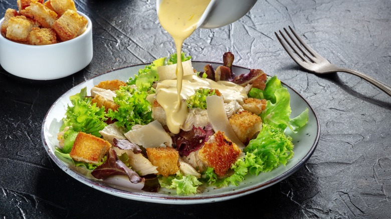 Yellow dressing pouring onto a plate of salad with a fork and a white ramekin of croutons