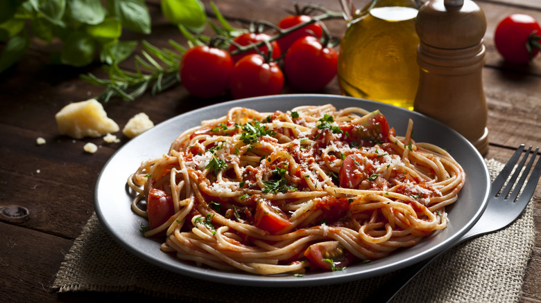 Spaghetti with red pasta sauce on a gray plate with tomatoes, basil, cheese, oil, and a pepper grinder behind it