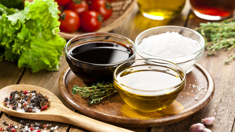 Small bowls of oil, vinegar, and salt sit on a round wooden tray surrounded by herbs, spices, and vegetables