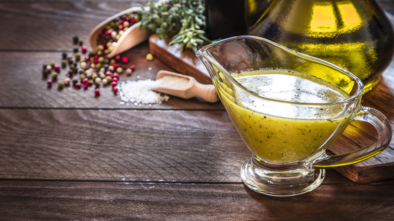 A small pitcher of vinaigrette sits on a wooden surface with salt and peppercorns in the background