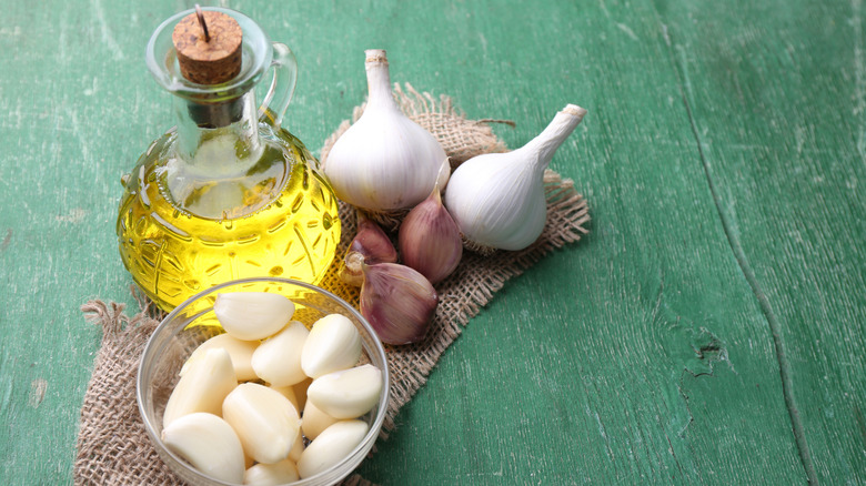 A bottle of olive oil and a bowl of whole, peeled garlic, surrounded by unpeeled garlic, on a piece of burlap on a green counter.