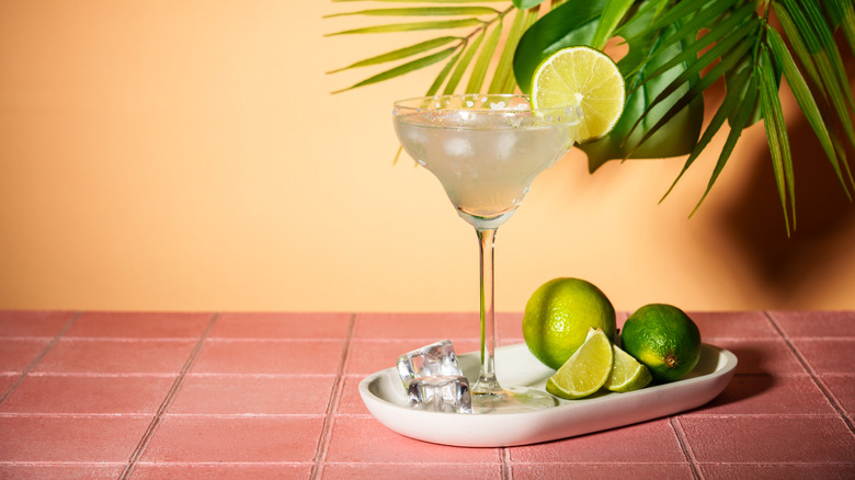 Margarita in a tall glass with lime wheel on a plate. Full limes and slices are on the plate with ice. All on a tiled table, with a light background and palms poking through the top right corner.