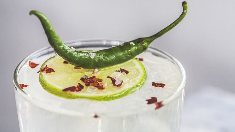 Close up of a margarita in clear glass, topped with lime wheel, chili pepper, and red pepper flakes on ice, with a white background.