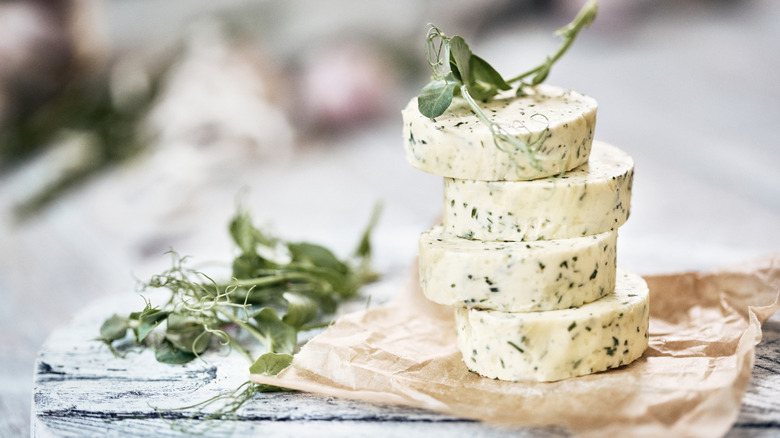 Garlic butter rounds stacked on parchment paper and light, wooden table with herbs on top and to the left.