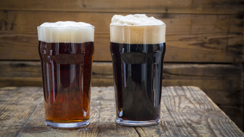 Two glasses of foamy beer, one reddish brown and one almost black, in pint glasses sit on a wooden background.