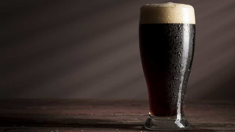 Dark beer with slight reddish tint and a foamy head in a tall glass sits on a wooden surface.