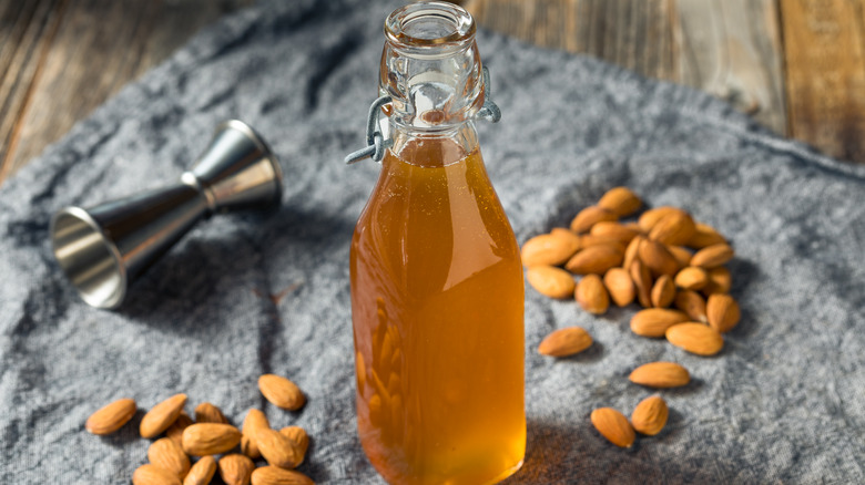 A bottle of homemade orgeat on a cloth napkin surrounded by almonds and a cocktail jigger