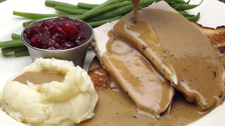 A plate of turkey, mashed potatoes, cranberry sauce and green beans with gravy being poured over the meat