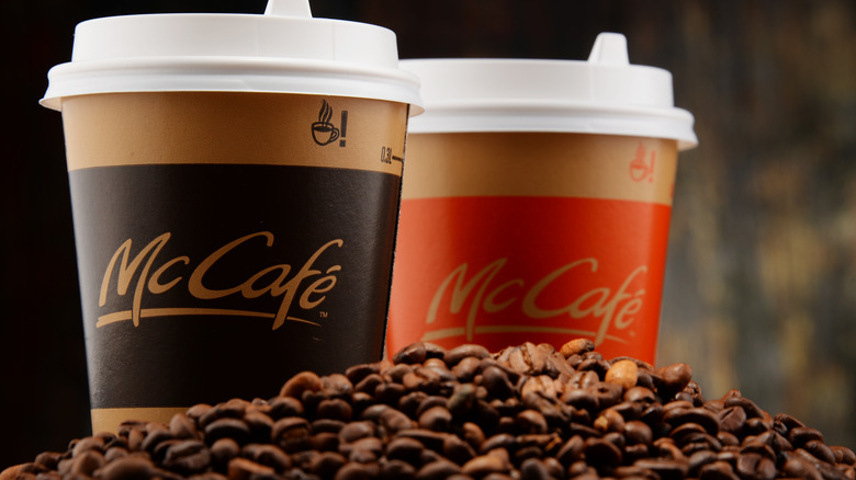 Two McCafe cups of coffee with white lids. Coffee beans are piled in front of the cups, with a blurred, texturized background.