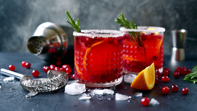 Two bright red colored cranberry margaritas with a sugared rim and rosemary sprig are surrounded by mixing equipment and garnishes