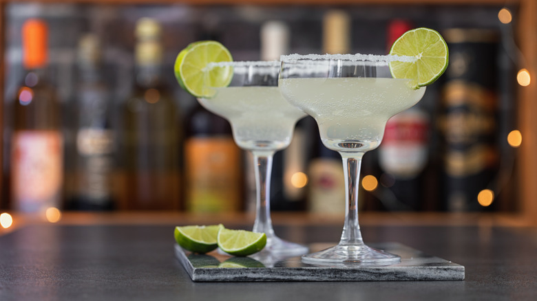 Two margarita glasses with a lime wedge and salted rim stand on a stone table