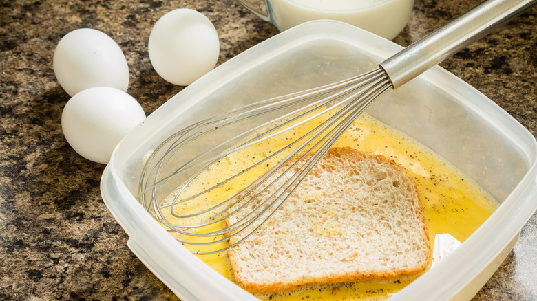 A piece of bread soaks in a French toast custard mixture inside a plastic dish, with a wire whisk also inside the dish and eggs and a measuring cup of milk sitting adjacent