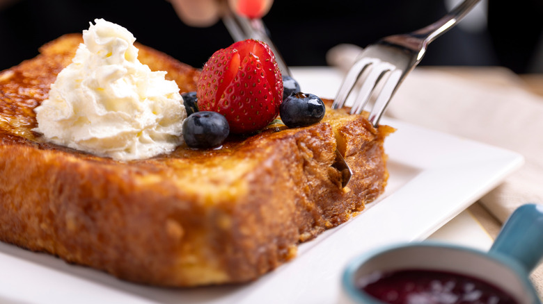 A knife and fork penetrate a piece of French toast on a white dish, topped with whipped cream and berries