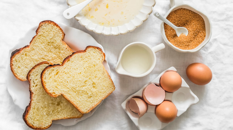 Ingredients for French toast, including a plate of bread, a small pitcher of milk, eggs, and a ramekin of cinnamon, sit on a white tablecloth