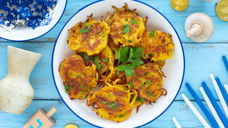 Plate of potato latkes on table with blue and white Hanukkah decorations, dreidels, gelt, and candles