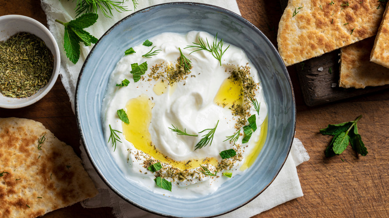 A bowl of labneh with oil, za'atar and herbs next to pita