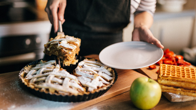 Person lifting a slice of pie to a small white plate in a kitchen, with an apple, waffles, and strawberries nearby, all on a wooden counter.