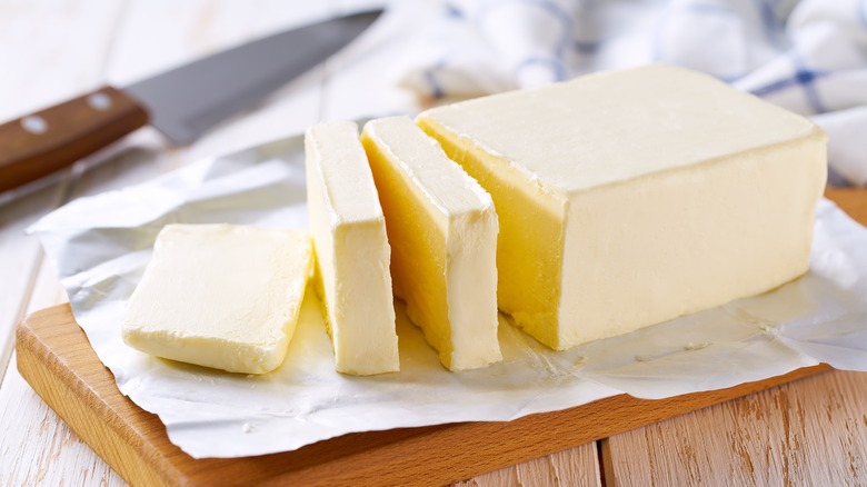 Close up of a stick of butter on a wooden cutting board with three cut slices and a knife in the background, all on a light wooden table.