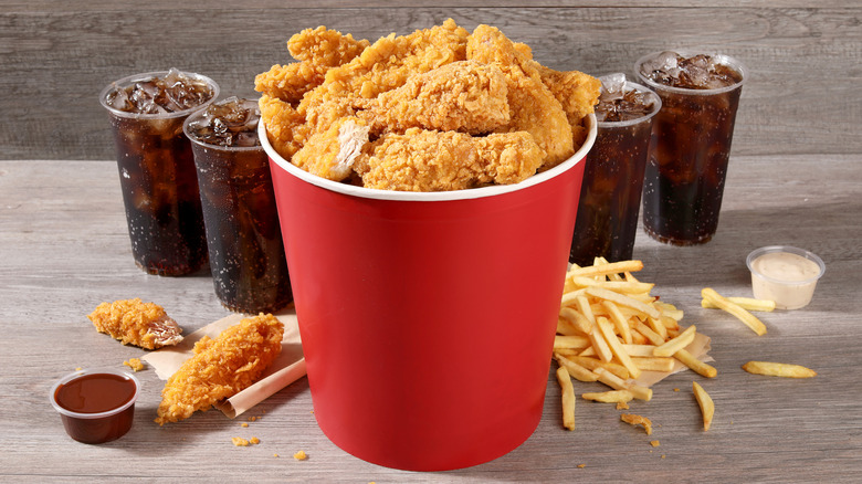 A red bucket of fried chicken surrounded by fries and soft drinks