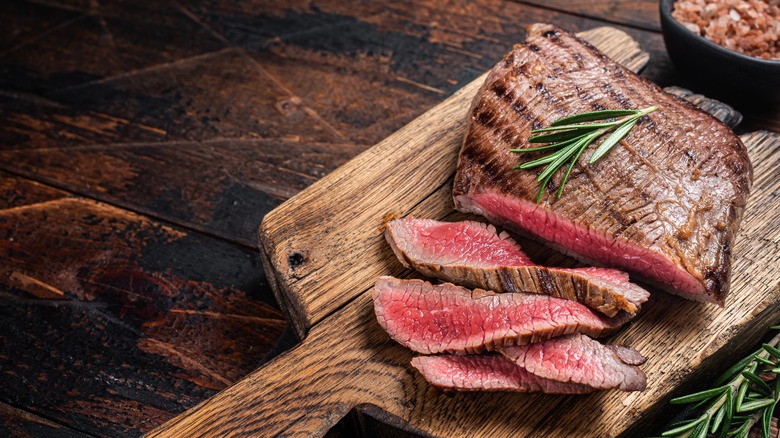 Flank steak topped with rosemary on a wooden cutting board on an aged wood table