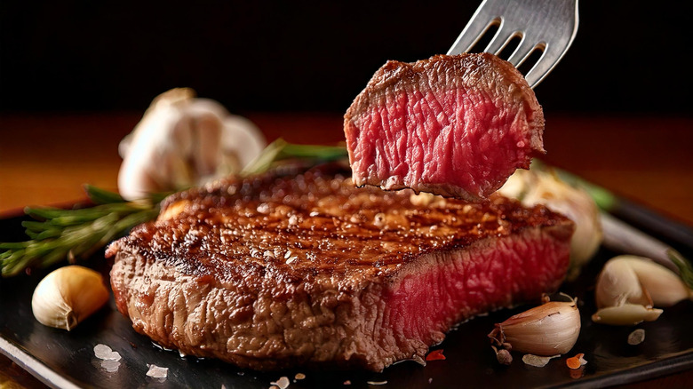 Medium rare steak on a black plate with a piece cut off that's on an elevated fork. The meat is surrounded by several cloves of garlic