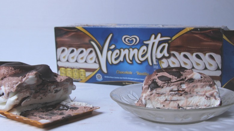 Slice of Viennetta ice cream cake on a glass plate, in front of product box. Small tray of Viennetta to the left with slight melt. All on a blank white table and background.