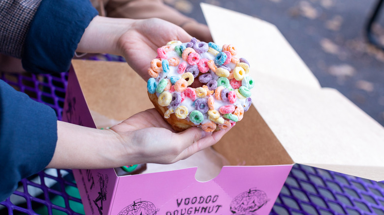 Two hands holding Voodoo Doughnut with fruit loop cereal on top over pink donut box on a purple outdoor bench