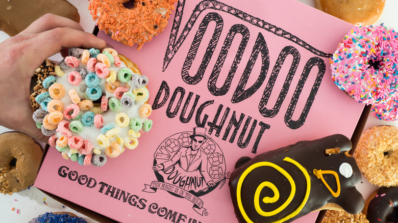 Voodoo Doughnut pink box with a variety of donut options surrounding and one is being held with a hand