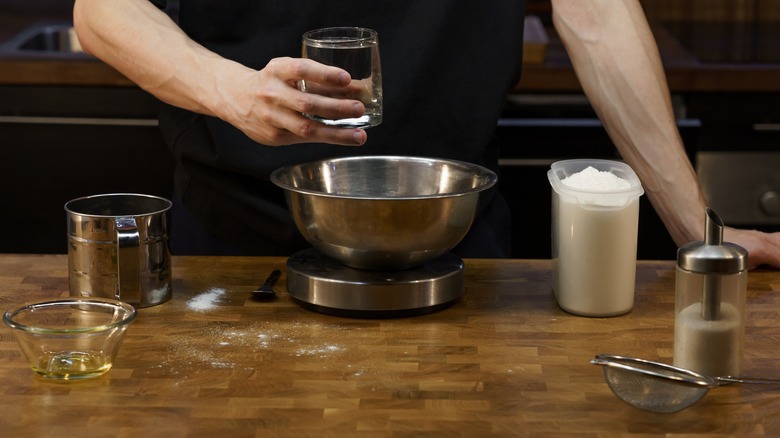 A cook puts pizza dough ingredients into a mixing bowl on a kitchen scale