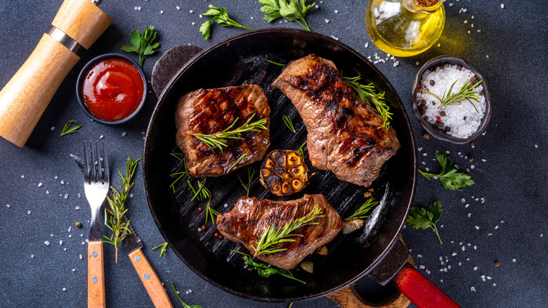 Three grilled steaks in a pan with oil, seasoning, and herbs