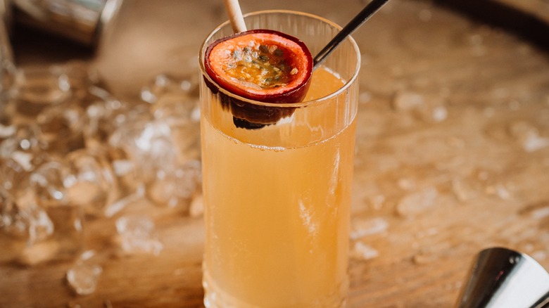 A light peach colored highball cocktail in a tall glass with a straw and half passion fruit garnish