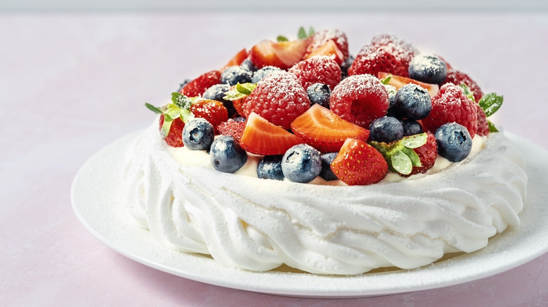 Round pavlova with a white base with whipped cream and strawberries and blueberries on top on a white plate