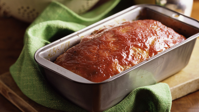 A glazed meatloaf in a pan rests beside a green kitchen towel