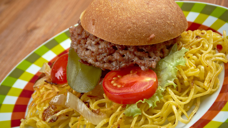 Chow Mein Sandwich with burger and a side of lettuce, pickle, and tomatoes sits on a checkered plate