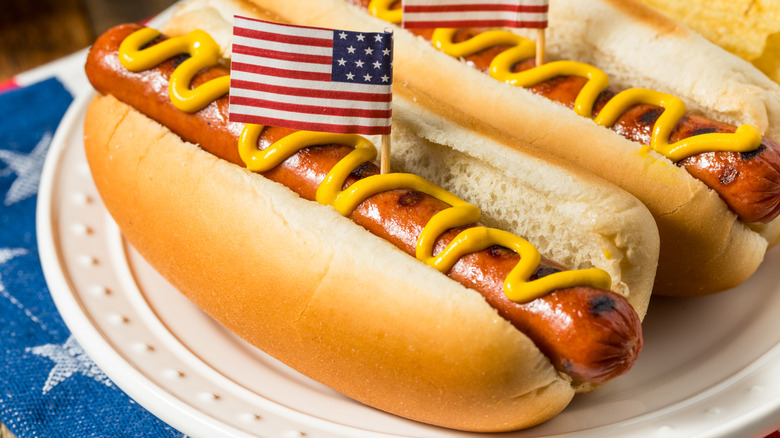 a hot dog in a bun with mustard and a tiny American flag on a plate