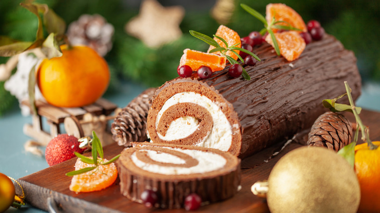 A yule log cake decorated for Christmas is topped with holly berries and slices of candied oranges