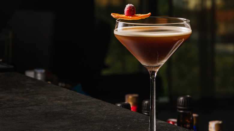 Dark brown cocktail in martini glass with raspberry and orange garnish